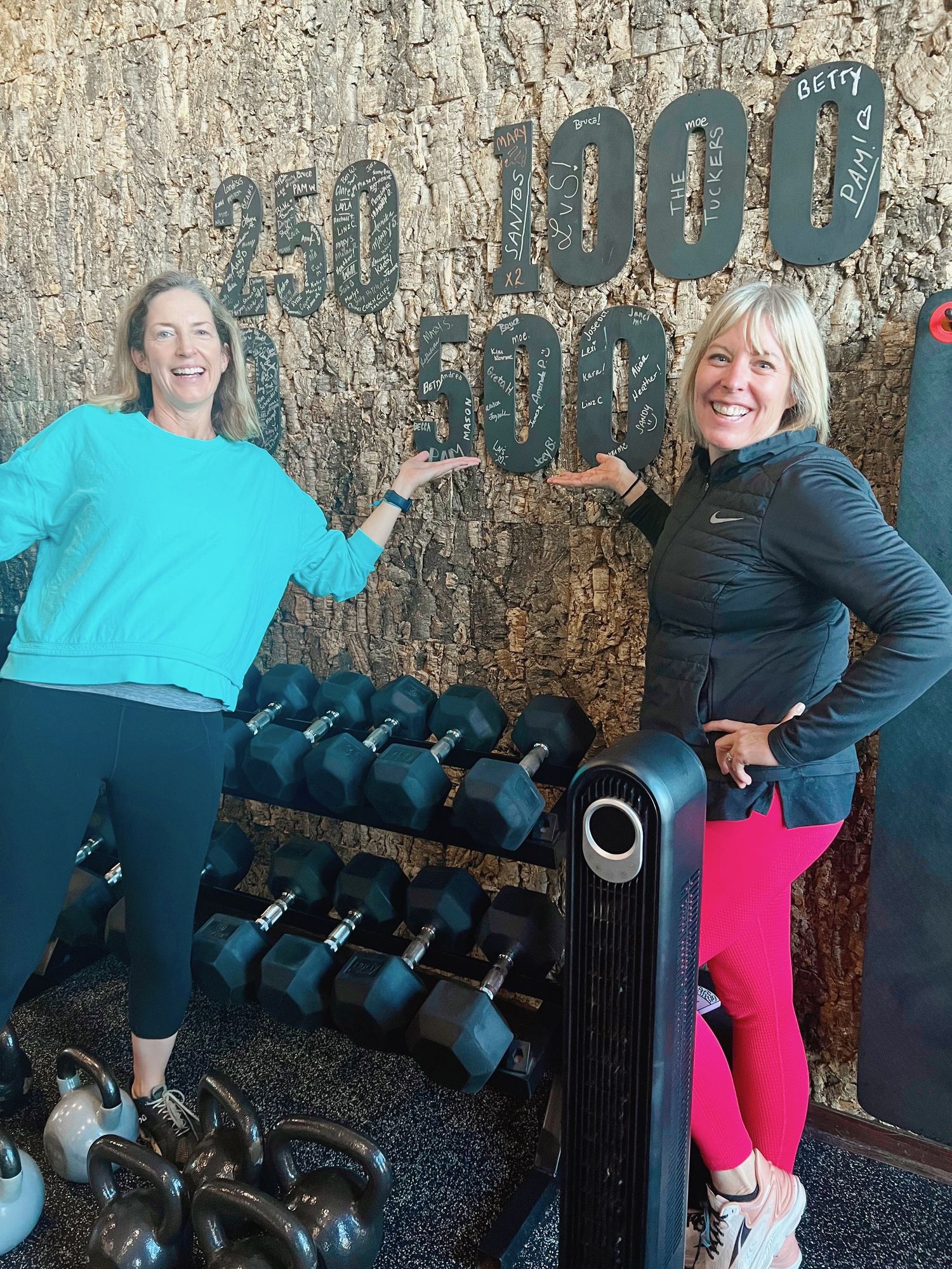Kara and a friend beaming at the gym, pointing at the signed 500-workouts milestone board