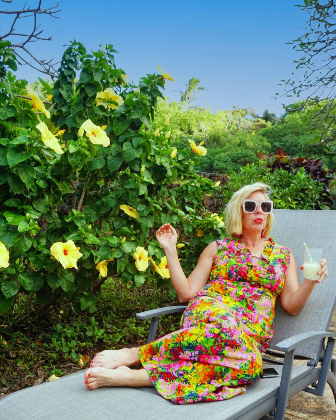 Kara reclining on a lounger in a bright floral dress with a drink, hibiscus behind her