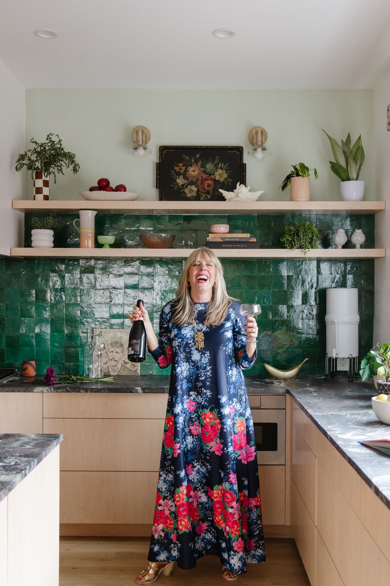 Kara laughing in her green-tile kitchen, bottle in one hand and a coupe glass in the other