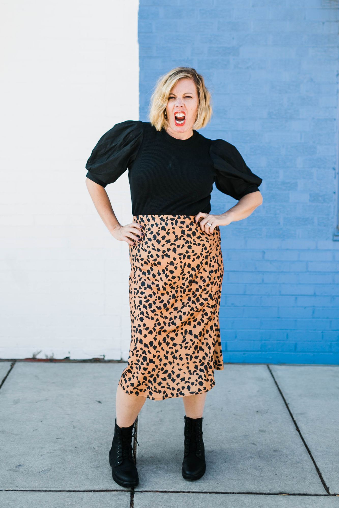 Kara yelling with joy in a leopard-print skirt and combat boots against a blue brick wall
