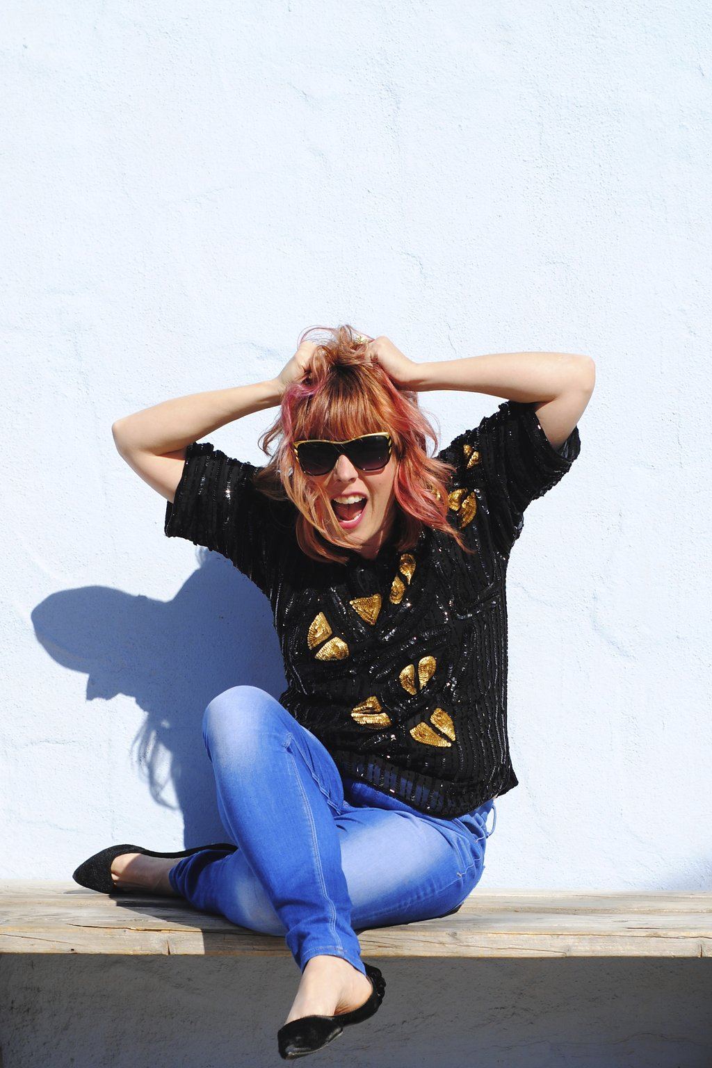 Kara, pink hair, sequin top, hands in her hair, laughing on a bench