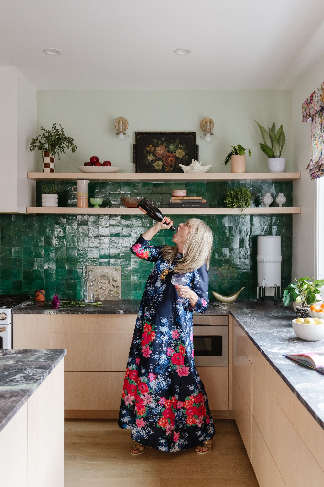 Kara in a floral caftan swigging from a bottle of bubbly in her green-tiled kitchen