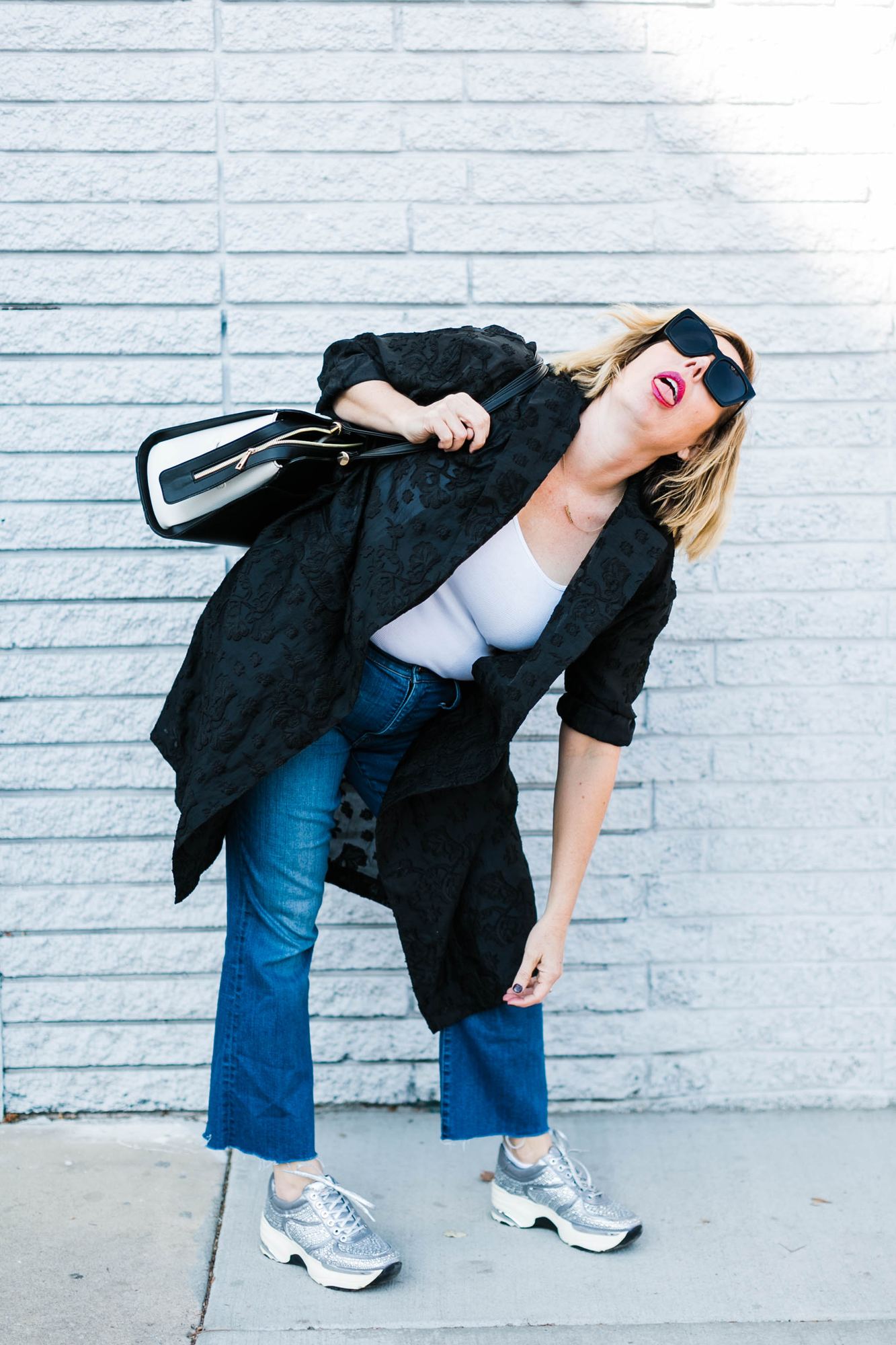 Kara bent over laughing against a white brick wall in a black duster and silver sneakers