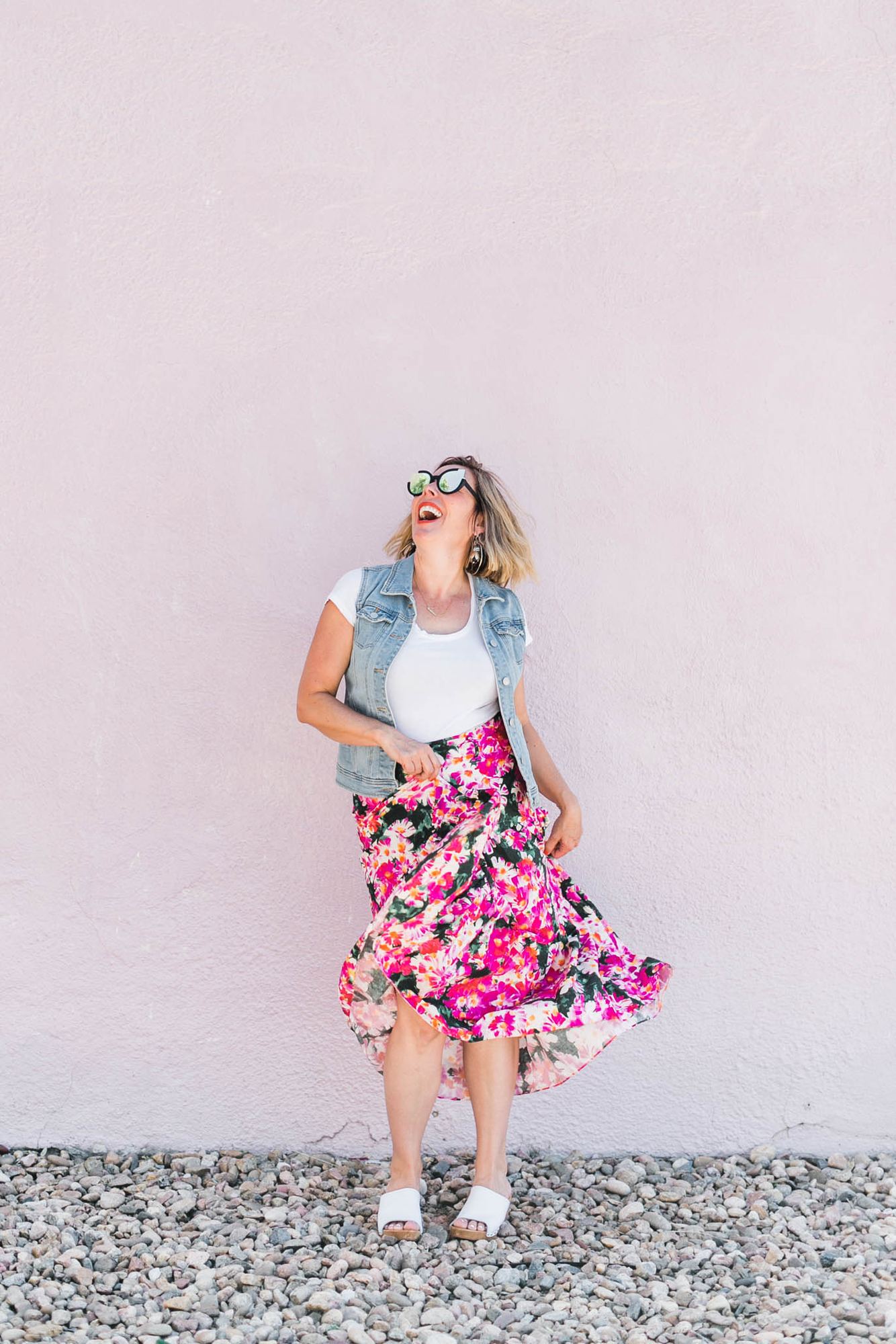 Kara laughing in sunglasses, a denim vest and a pink floral skirt against a pink wall