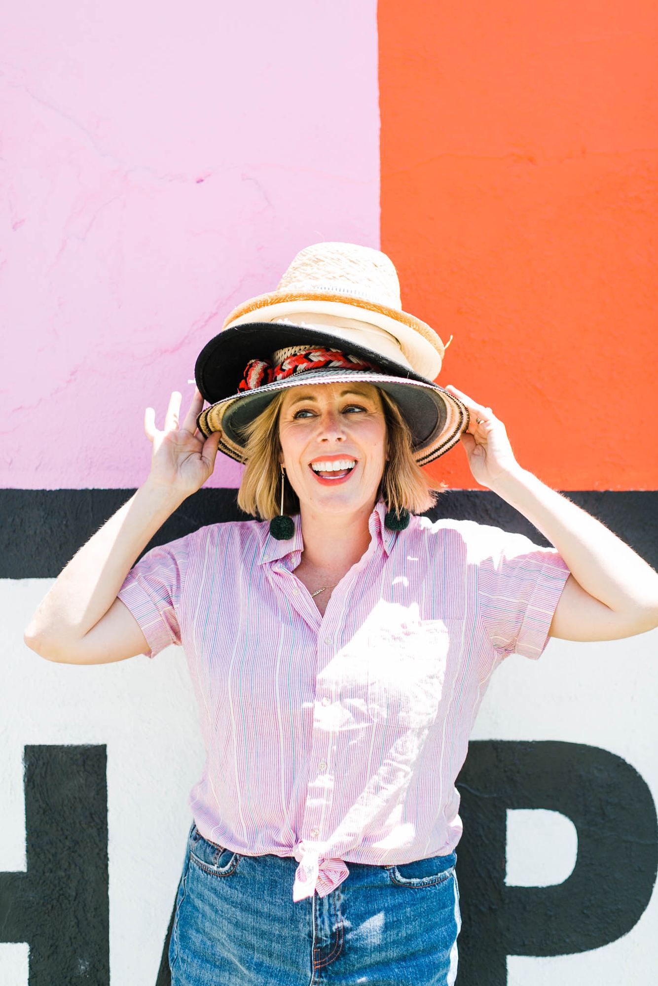 Kara laughing under a stack of six hats against a pink and coral wall