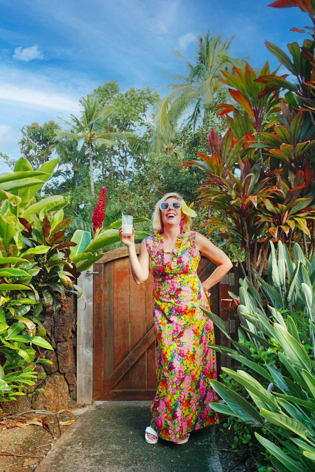 Kara laughing with a drink in a bright floral maxi by a garden gate