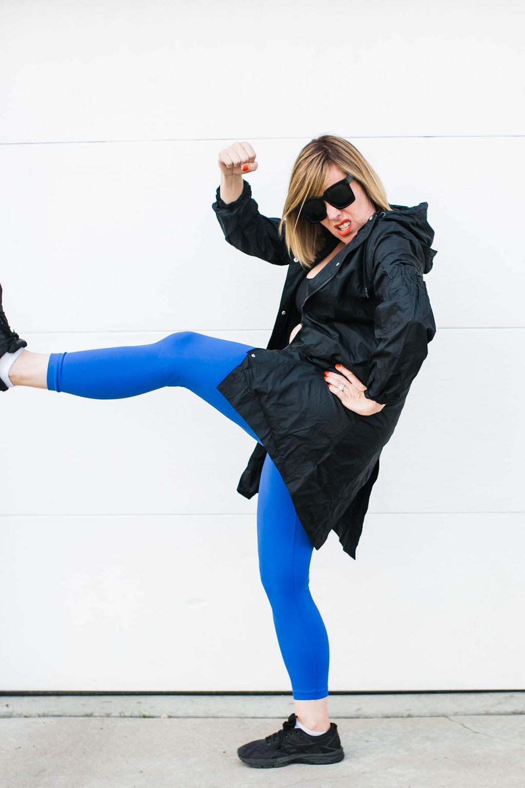 Kara in a black coat and blue leggings, karate-kicking in sunglasses