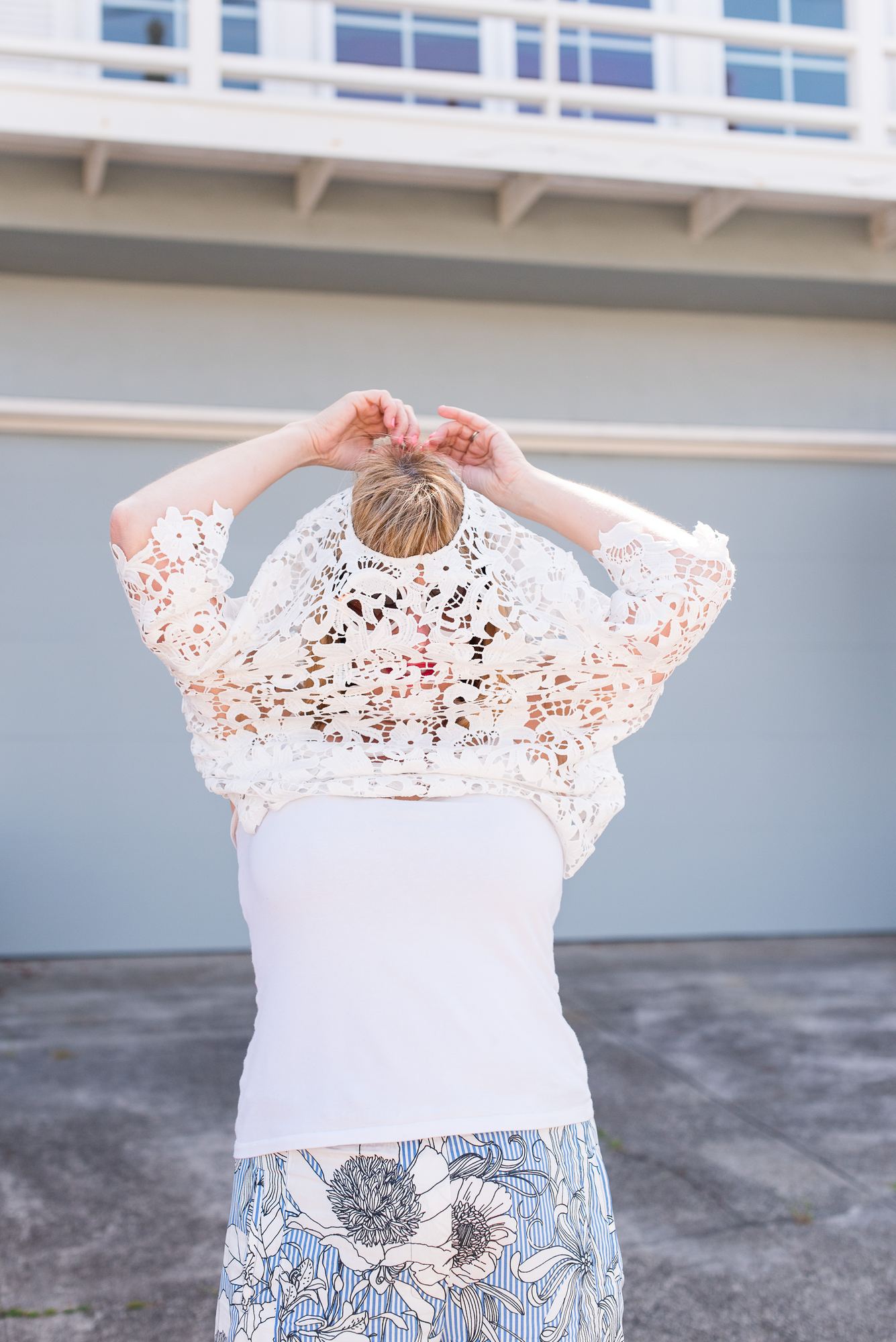 Kara from behind, pulling a white lace top up over her head