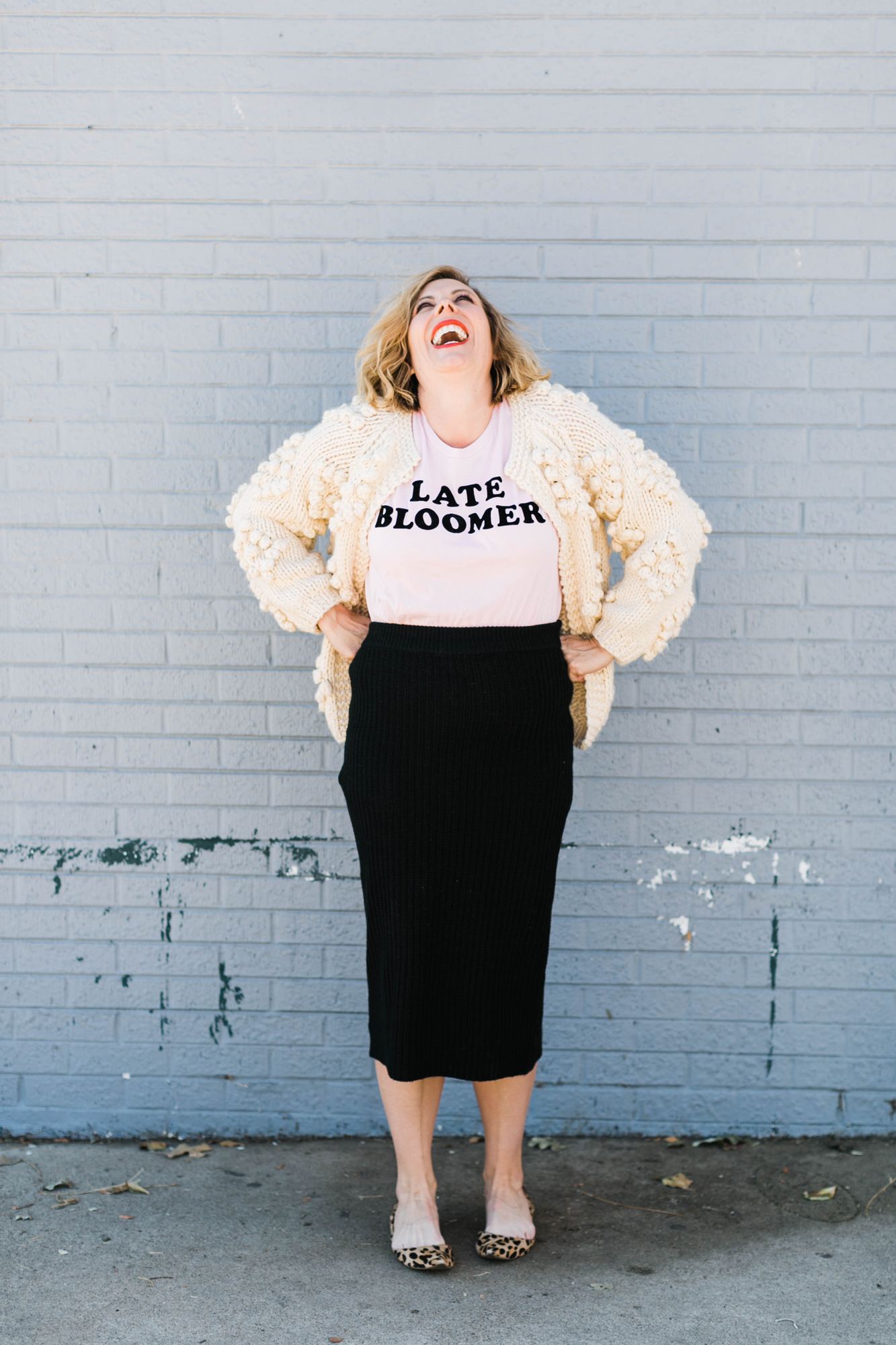 Kara laughing up at the sky in a Late Bloomer tee, cream bobble cardigan and leopard flats