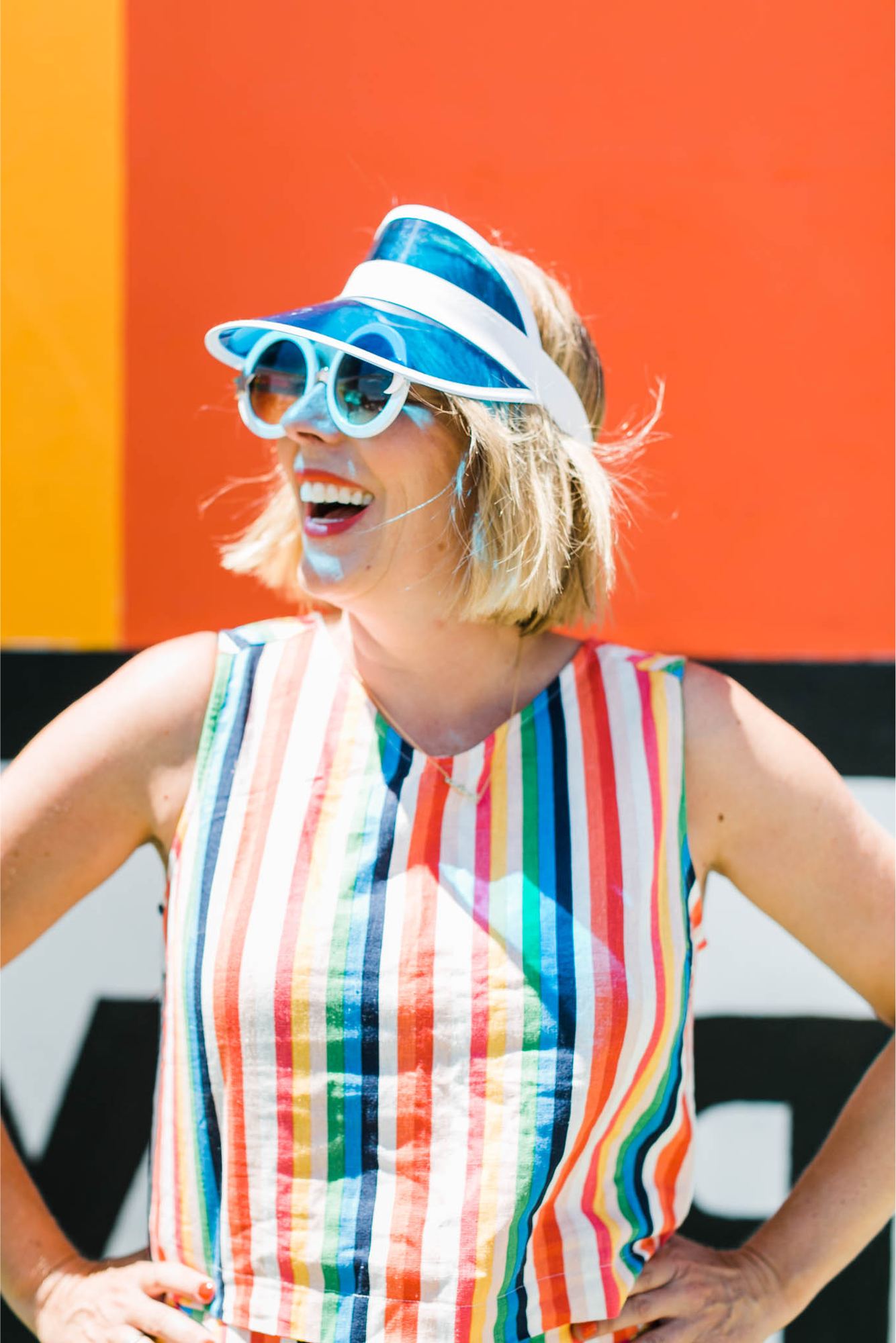 Kara laughing in a rainbow-striped top, blue visor and round sunglasses against an orange wall